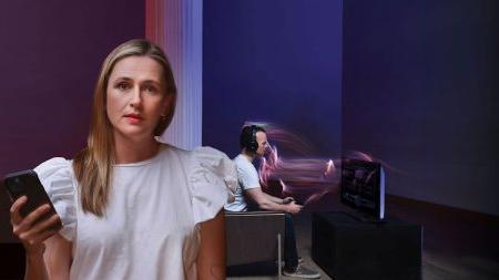 前景中的女人带着关切的神情看着镜头, 而她身后的男人在玩电子游戏.